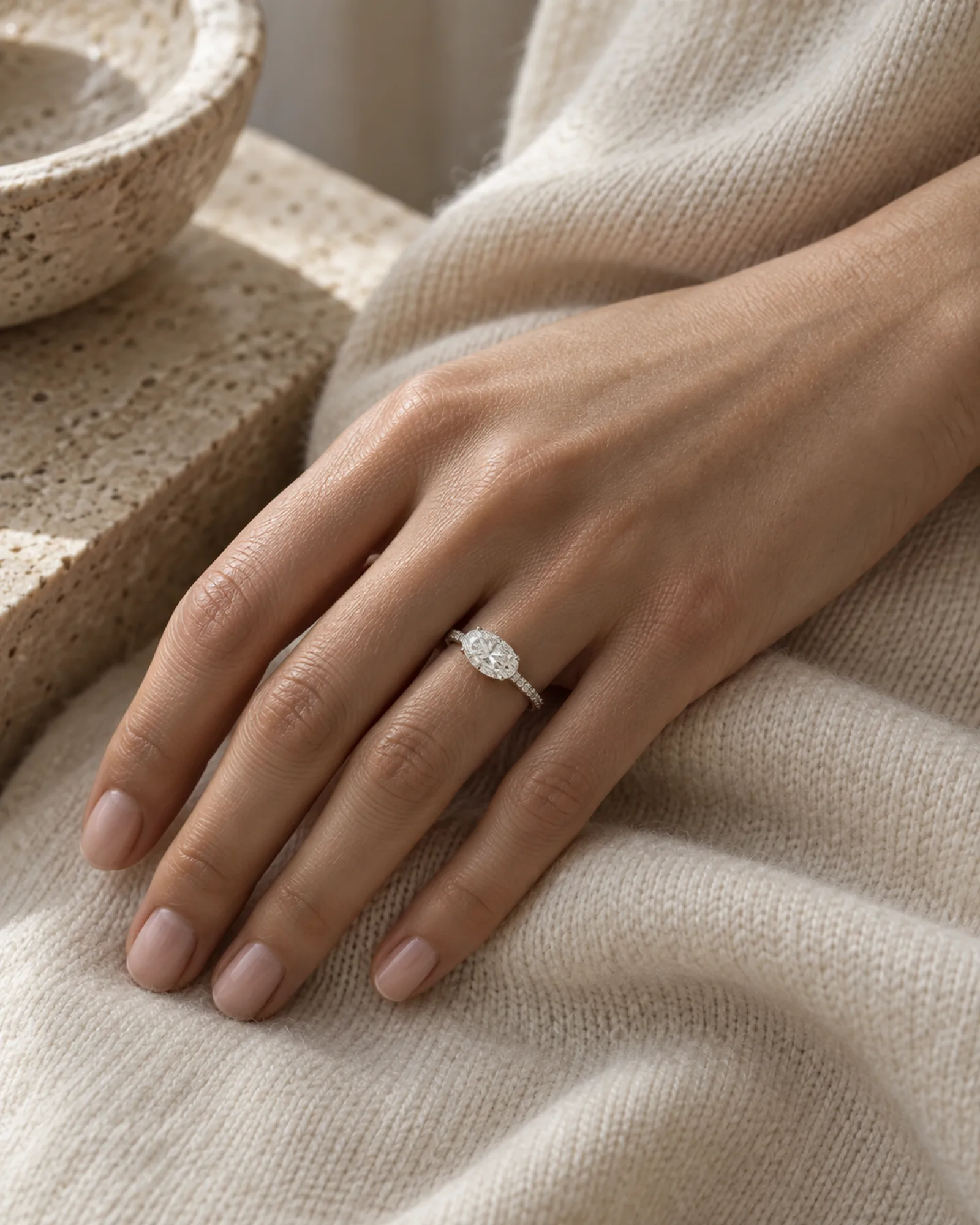 A hand resting on ivory cashmere wearing a platinum ring with an east-west oval diamond and a thin pavé band